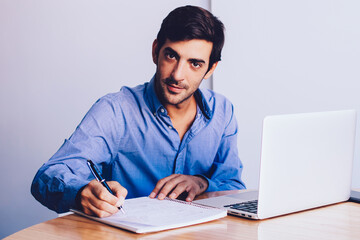 Portrait of young entrepreneur sitting at desk with laptop computer and writing main theses for business deal, indian director looking at camera enjoying paperwork at modern office with good wifi