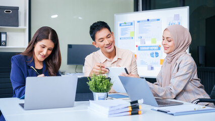 A diverse group of professionals, including a male Asian businessman and a female Muslim businesswoman in hijab, gather around a desk equipped with tablets and laptops for a productive meeting.