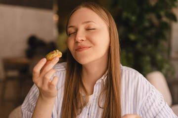 Young blonde woman eating eclair sitting in cafe. Girl bite piece of croissant look joyful at...