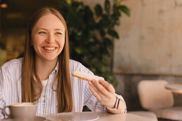 Young blonde woman eating eclair sitting in cafe. Girl bite piece of croissant look joyful at...