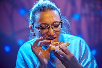 An adult woman 45 years old wearing glasses enjoys eating pizza during her lunch break.