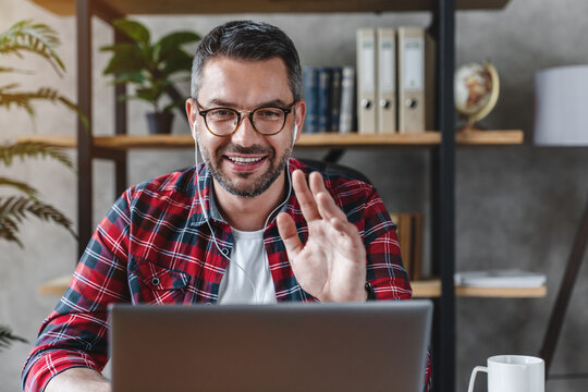 Front shot of smiling businessman freelancer entrepreneur sitting a home office looking a laptop screen having online meeting work discussion conference video zoom call, waves his hand and says hi