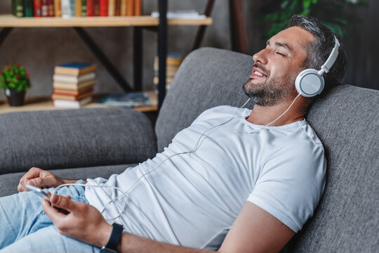 Joyful caucasian man at home lying resting on sofa listening music e-book podcast song using holding smartphone mobile phone wearing white headset earphones . Time to relax.