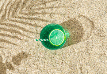 Creative sunlight summer scene of green refreshing cocktail drink with lime slice and tropical palm...