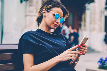 Charming teenage hipster girl sitting at bench and using mobile phone for blogging, young pretty...