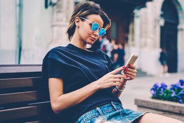 Charming teenage hipster girl sitting at bench and using mobile phone for blogging, young pretty...