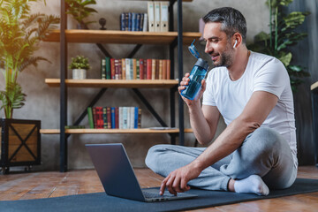 Adult man using laptop after online fitness lesson workout holding bottle drinking water with...