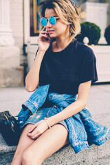 Charming woman with mobile phone calling to friend using application while recreating on urban setting outdoors, hipster girl wear in casual denim look talking by advanced telephone outdoors