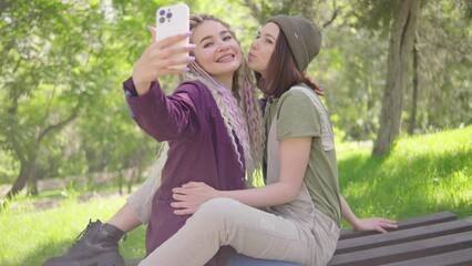 Young adult lesbian couple sitting face to face on bench and taking selfie in sunny park - Powered by Adobe