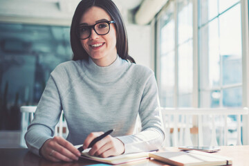Smiling brunette hipster girl wear in fashionable spectacles studying at coworking space and...