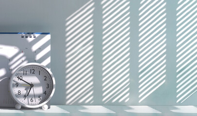 White round table clock and desk calendar on tabletop with morning sunlight and shadow on surface,...