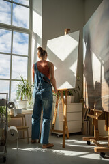 Artistic woman painter at easel, clean blank canvas in light sunny art studio. Beginning of working day, positive optimistic atmosphere of creative workshop. Rear back view of girl preparing to work.