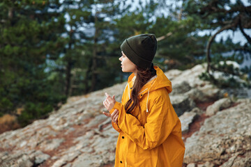 A woman in a yellow raincoat standing on a rocky hill with trees in the background, enjoying nature and adventure