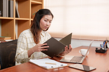 Young Asian woman reading folder while sitting at desk with laptop, documents, and cup of coffee....