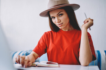 Portrait of attractive concentrated woman in trendy titfer looking at camera and working remotely with computer, beautiful female freelancer in stylish apparel planning distance job in cafeteria