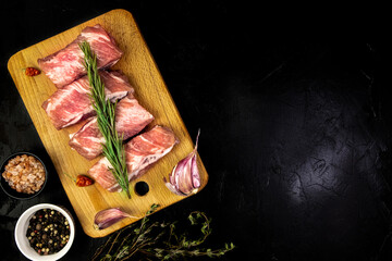 Raw seasoned pork belly on a cutting board with herbs on a black concrete background, top view,...