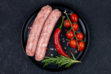 Raw meat sausages with herbs and tomatoes on a black plate close-up on a black concrete background