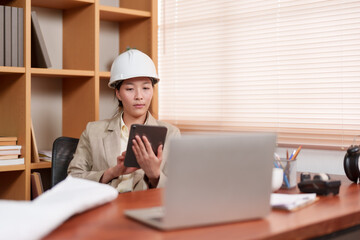 Young Asian woman in hard hat and suit using tablet at desk with laptop. Office environment with bookshelves, documents, plants. Focused on work, representing an architect or designer.