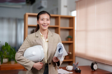 Asian female architect holding white hard hat and rolled blueprints, standing in modern office....