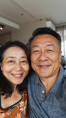 A man and woman smiling at the camera standing close together with the man wearing a blue shirt and the woman in a patterned top set against a blurred indoor background.