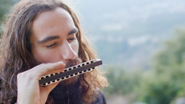 musician enjoys playing the harmonica while standing on the street. a young man with long hair plays the harmonica.