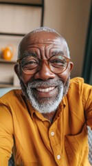 Obraz premium Smiling elderly man with glasses and white beard wearing yellow shirt sitting in a room with a shelf in the background.
