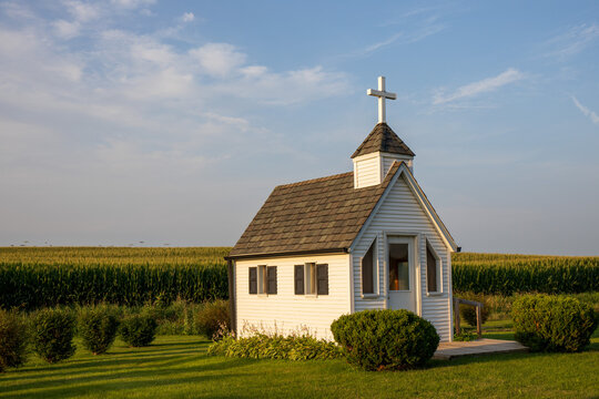 Tiny church on the edge of a corn field with grass in the foreground.  White church under blue sky with light clouds.  Tiny chapel in rural Iowa. 