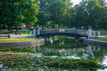 Fototapeta premium Green trees and pond with reflective surface. Pedestrian bridge over clear smooth pond. Central College campus in Pella, Iowa. 