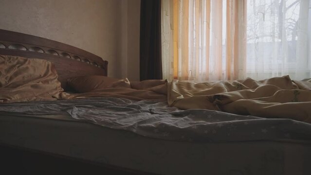 Unmade bed with rumpled sheets in softly lit room. Morning light streams through sheer curtains casting warm glow.