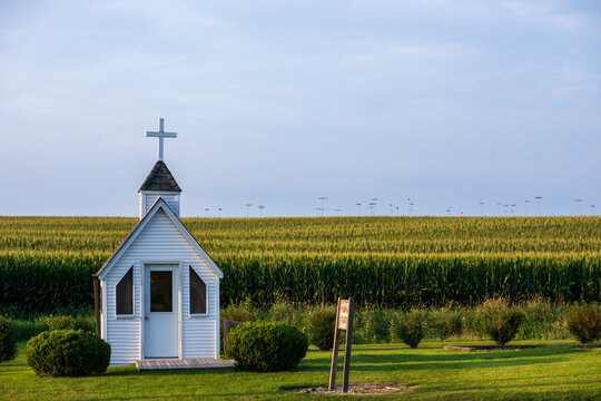 Tiny church on the edge of a corn field with grass in the foreground.  White church under blue sky with light clouds.  Tiny chapel in rural Iowa. 