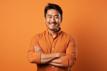 Fototapeta premium Portrait of a satisfied asian man in his 30s with arms crossed isolated on pastel orange background