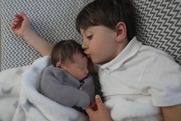 Tender moment of an older brother gently kissing his newborn sibling while they rest on a patterned blanket.