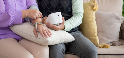 Closeup hands asian senior couple sitting on sofa checking blood pressure with pressure gauge in living room at home, elderly man and woman checkup health and pressure, medical of patient.