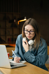 Portrait of student woman sitting at desk, using laptop and writing in notebook.