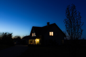 Warm lights glow from the windows of a house under the twilight sky, casting a serene ambiance over...