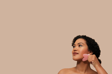 Young woman with curly hair performs a facial massage using a pink gua sha stone, a traditional Chinese medicine tool. She is captured mid-movement with the stone held against her jawline, copy space