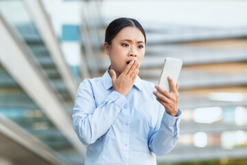 Fototapeta premium A young Asian woman dressed in a light blue shirt expresses shock as she reads a message on her smartphone. She is standing outdoors with an urban backdrop