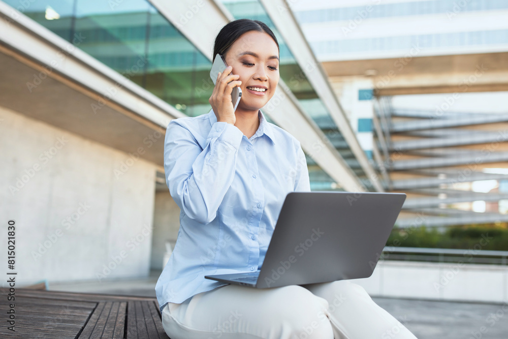 Poster Asian woman is seated on a bench, engrossed in a phone conversation while simultaneously working on a laptop. She appears focused and multitasking efficiently in a public setting.