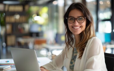 Smiling woman in office, using laptop, wearing glasses.