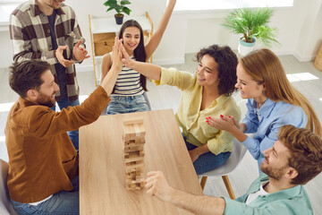 Portrait of excited happy young friends guys and girls playing together in board game with wooden...