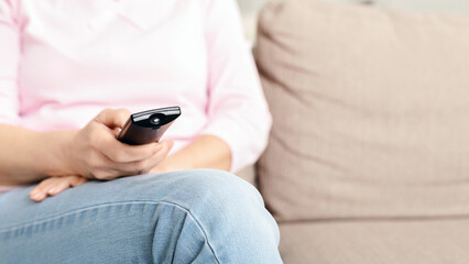 Cropped of woman is seated comfortably on a couch, holding a remote control in her hand. She...