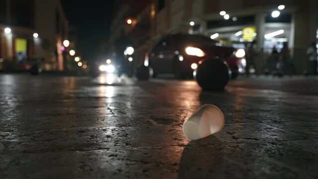 Close up of an empty plastic cup on the sidewalk at night illuminated by the headlights of passing cars. People throwing trash on the city street