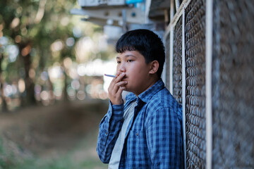 Teenager underage smoking a cigarette causes bad habits causing addiction. Asian kid smoking behind the school, an old dirty grungy place.