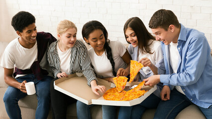 A diverse group of teenagers is sitting together, smiling and enjoying a large pizza. They are casually dressed, indicating a relaxed and social atmosphere