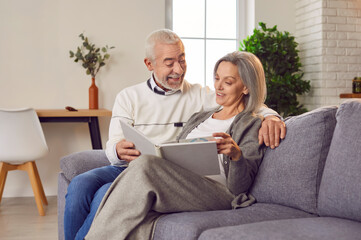 Joyful aged couple sitting on sofa looking their family photo album indulging in happy memories....