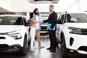 A professional car salesman, dressed in a suit, is shaking hands with a happy Indian couple...