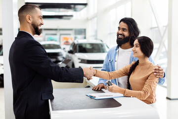 A man and a woman are shaking hands inside a well-lit car showroom. The man appears to be a car...