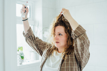 Haircare. Young plus size Lady Applying Hair Spray For Curls While Standing Near Mirror In...