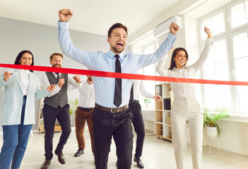 Businessman running and crosses the red finish line in the office, with colleagues supporting and...