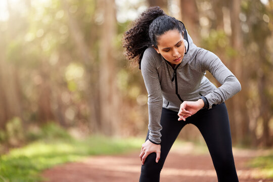 Woman, tired and checking watch in nature for distance, fitness and wellness in forest. Brazilian runner, taking break and counting steps from workout, challenge and goals in woods with copy space